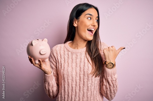 Young beautiful brunette woman holding piggy bank saving money for retirement pointing and showing with thumb up to the side with happy face smiling