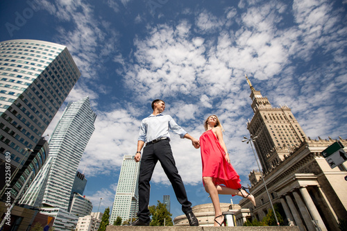 Bottom view of a young romantic couple. Woman in red dress walks holding hands with a man on the background of the city and blue sky with clouds. Poland, Warsaw