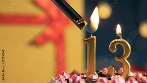 Birthday cake number 13 golden candles burning by lighter, background gift yellow box tied up with red ribbon. Close-up