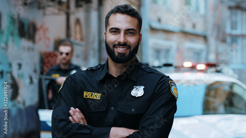 Smiling young man cops stand near patrol car look at camera enforcement happy officer police uniform auto safety security communication control policeman portrait close up slow motion