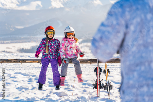 Little girls in outwear and masks with helmet sitting on wood fence with skis near against mountains