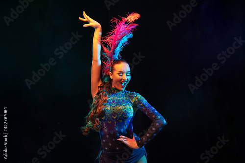 Rhythm. Beautiful young woman in carnival, stylish masquerade costume with feathers on black background in neon light. Copyspace for ad. Holidays celebration, dancing, fashion. Festive time, party.