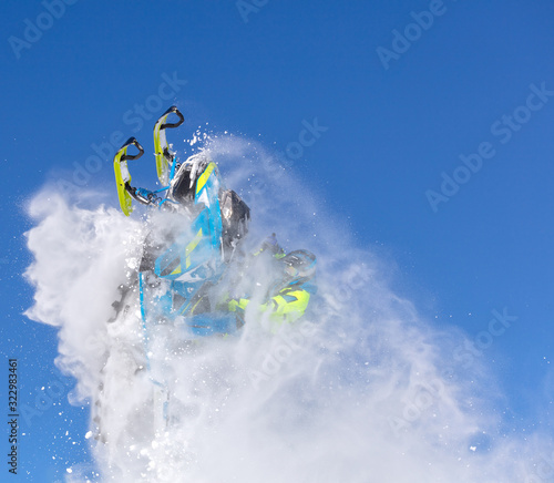 elite sports snowmobiler rides and jumps on steep mountain slope with swirls of snow storm. background of blue sky leaving a trail of splashes of white snow. bright snowmobile and suit without brands
