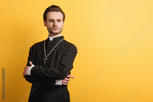 confident catholic priest standing with crossed arms and looking away isolated on yellow