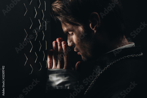young catholic priest praying with closed eyes near confessional grille in dark with rays of light
