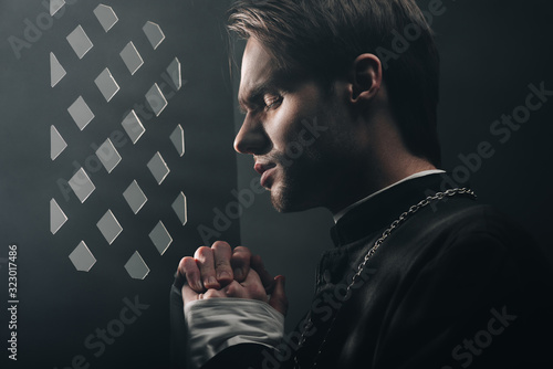 young thoughtful catholic priest praying with closed eyes in dark near confessional grille with rays of light