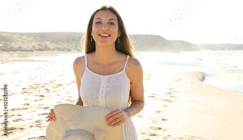 Portrait of beautiful smiling carefree woman walking on the beach in white dress holding straw hat looking at camera with overexposed background. Copy space.