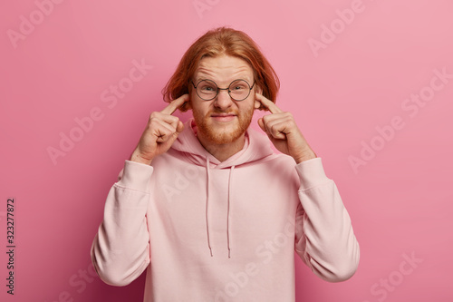 Image of stressed redhaired man bothered by loud neighbour, tells turn off music, unwilling hear awful sound, plugs ears, smirks face, wears spectacles and hoodie, poses indoor over rosy wall