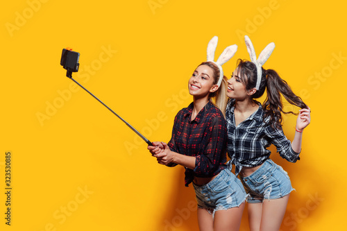 Two Young sexy caucasian woman wearing cute easter rabbit ears making selfie using stick over yellow isolated background.