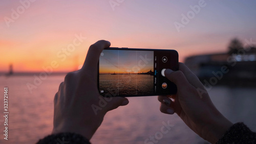 Close-up of a woman's hands with a phone taking photos of a bright sunset and the sea.