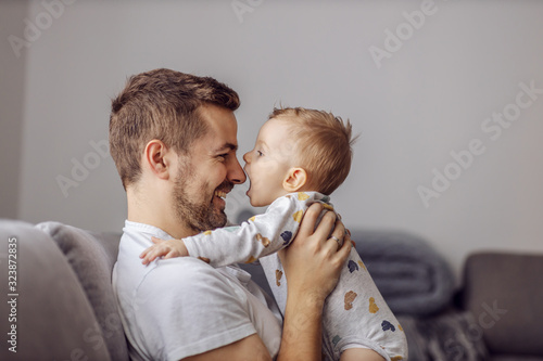 Adorable little blond boy playing with his caring father and trying to bite his nose. Family values.