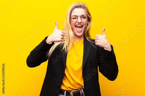 young pretty blonde woman smiling broadly looking happy, positive, confident and successful, with both thumbs up against yellow wall