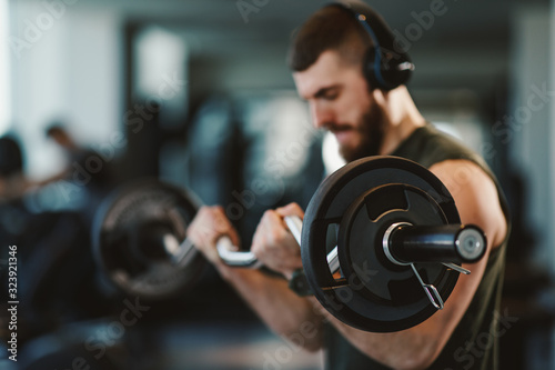 Young bearded man doing biceps exercises in gym with barbell selective focus on weights