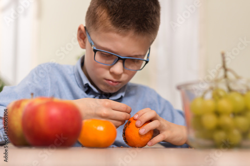 Chłopiec w okularach ze skupieniem wbija w owoc mandarynki goździki. Zdobienie owoców mandarynki goździkami przez chłopca w wieku szkolnym. Na pierwszym planie na blacie leżą owoce jabłka i mandarynki