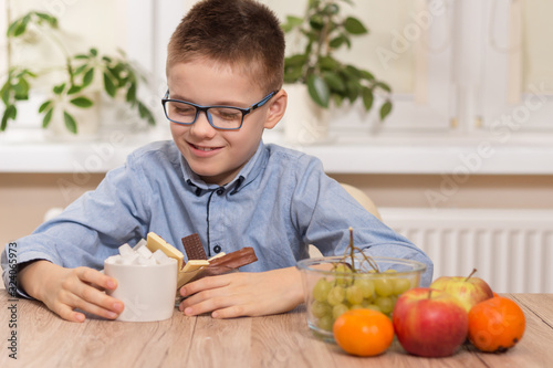 Chłopiec w wieku szkolnym z uśmiechem wpatruje się i trzyma naczynie ze słodyczami oraz cukiernicę z kostkami cukru. Obok na blacie stołu stoją owoce mandarynki, winogrona i jabłka.