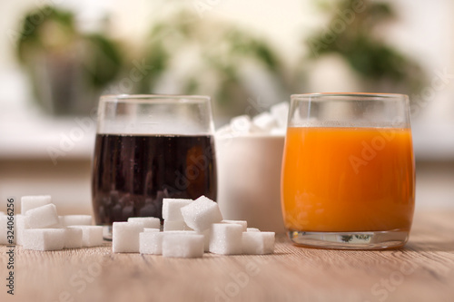 Kostki cukru porozrzucane leżą na blacie kuchennym. W jednej szklance stoi cola a w drugiej sok. Na drugim planie cukiernica wypełniona kostkami cukru.