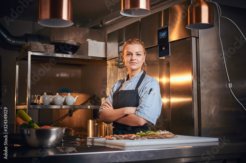 Confident and serious female chef standing with hands crossed in a dark kitchen next to cutting board with vegetables on it, wearing apron and denim shirt, looking in the camera, cooking show look