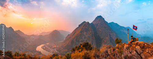 Sunset panoramic view of couple of trekkers sitting on a rock on top of Nong Khiaw View Point with beautiful mountain and Nam Ou river in background, Luang Prabang Province, Northern Laos.