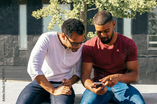 Excited male friends studying mobile app features on phone. Two men in casual sitting on parapet outside, using one smartphone and staring at screen. Mobile app concept