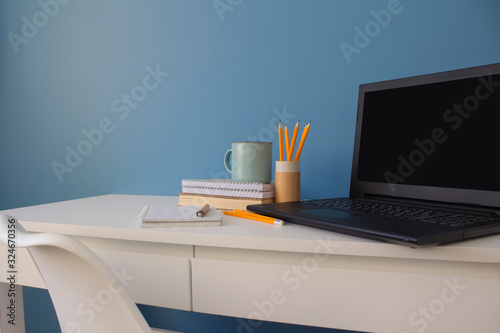 Empty home office of creative entrepreneur with black laptop on white table during lunch break. Modern workspace with blue cup of coffee and yellow pencils in paper tube as reusable packaging concept.