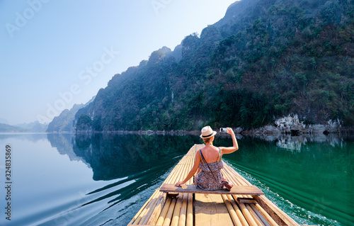 Travel and technology. Pretty young woman taking photo on smartphone sailing National Park lake on traditional bamboo raft.
