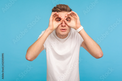 Portrait of curious man in casual white t-shirt looking through fingers in binoculars gesture, observing distant with attentive look, watching afar. indoor studio shot isolated on blue background