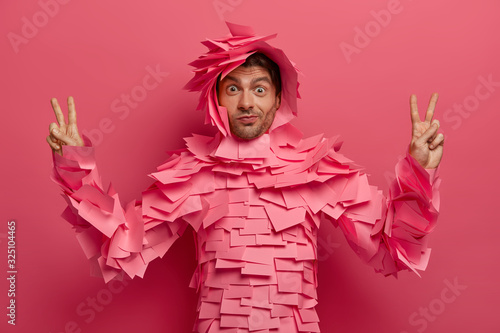Surprised funny man has fun in office, poses in creative costume made of sticky notes, raises fingers in victory gesture, shows peace sign, isolated over pink background. Paper outfit. Monochrome