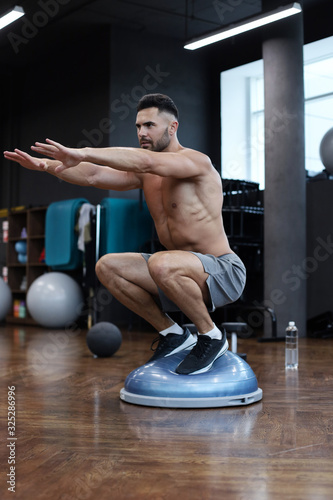Fit athlete performing exercise on gymnastic hemisphere bosu ball in gym.