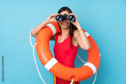 Lifeguard woman over isolated blue background with lifeguard equipment and with binoculars
