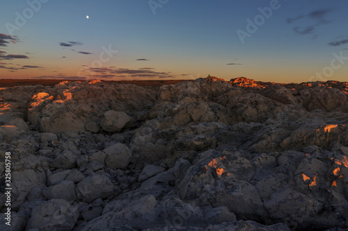 Many gray stones with sharp edges are lit by the rays of the setting sun. The moon and stars in the blue evening sky. Traveled photo in Mongolia.