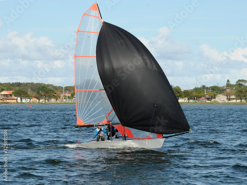 Kids racing a small sailboat with a Black spinnaker at a Junior yachting regatta on a coastal lake.