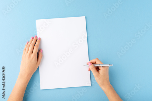 Overhead shot of female hands writing with pen over empty white sheet of paper on blue background