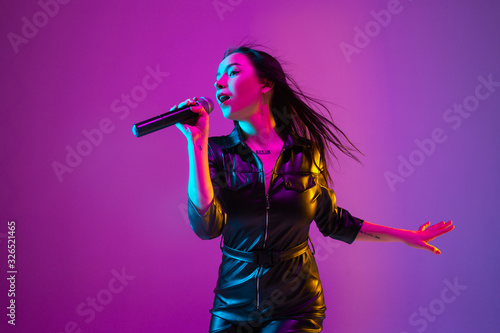 Caucasian female singer portrait isolated on purple studio background in neon light. Beautiful female model in black wear with microphone. Concept of human emotions, facial expression, ad, music, art.