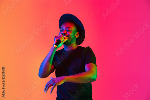 Young african-american musician singing on gradient orange-purple studio background in neon light. Concept of music, hobby, festival. Joyful party host, stand upper. Colorful portrait of artist.