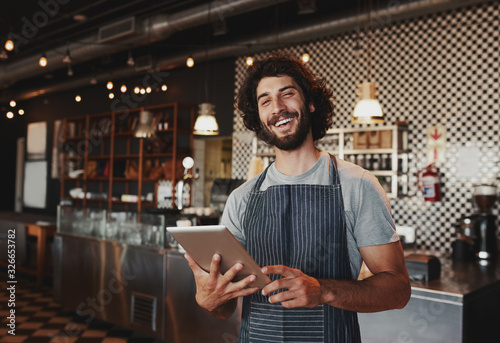 Handsome young caucasian coffee shop owner wearing apron laughing using digital tablet