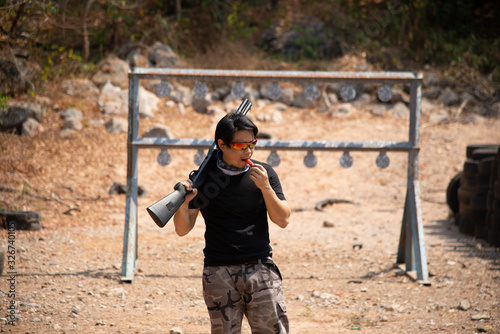 military sport man short hair wear solid black shirt, camoflage plant, camouflage scarf and red dark shade holding black shotgun on right shoulder and loking on left standing in shooting range