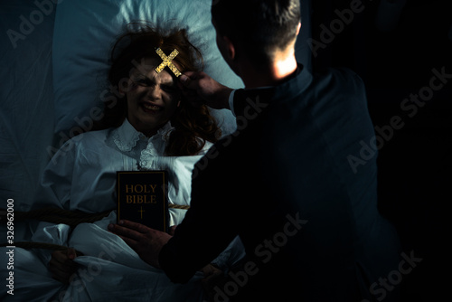 exorcist with bible and cross standing over demonic obsessed girl in bed
