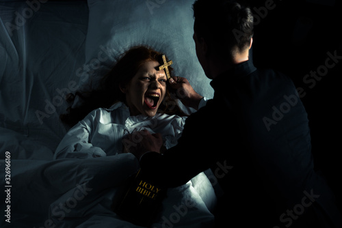 exorcist with bible and cross standing over demoniacal yelling girl in bed