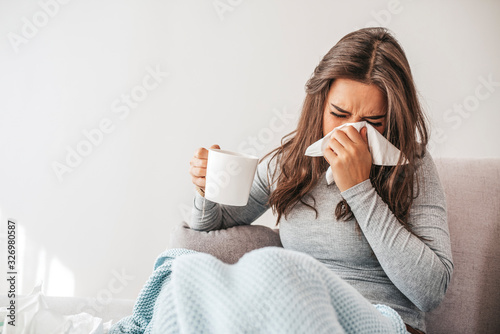 Sick woman covered with a blanket lying in bed with high fever and a flu, blowing her nose. Pills and glass of water on the table. Woman sneezing in a tissue in the living room