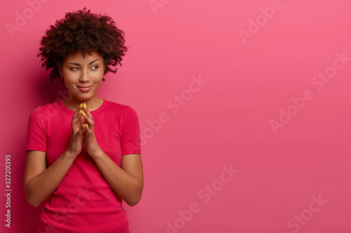 Pretty woman steepls fingers, looks with devious expression aside, has intention, thinks over brilliant plan, has charming smile, wears casual rosy t shirt, isolated over pink background, empty space