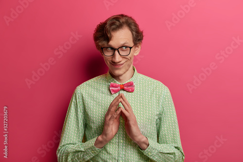 Isolated shot of cunning thoughtful man has evil genius plan, steepls fingers with intention to do something, has pleased pensive expression, wears elegant green shirt and bow tie, stands indoor