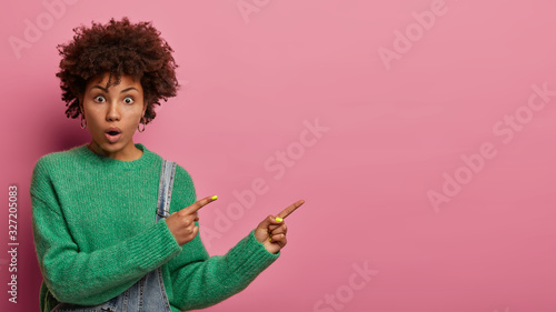 Photo of surprised curly haired young woman indicates away, shows free space for your promotion, fascinated by high prices, gasps from wonder, wears green sweater, isolated on pink background