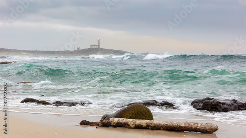 A tree trunk lies on the shore in the north of Spain in Galicia on the sand in front of a stone. In the background the Larino lighthouse on the Atlantic coast. Big waves on the beach.