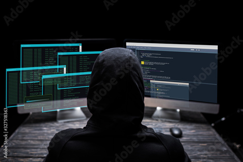 back view of hooded hacker sitting near computer monitors with data on screens on black