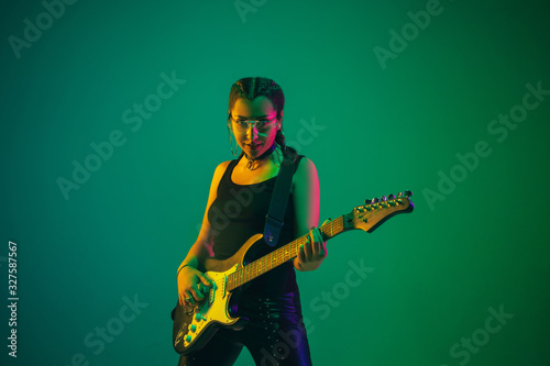 Rockstar. Caucasian female guitarist portrait on green studio background in neon light. Beautiful female model in black wear with guitar. Concept of human emotions, facial expression, ad, music, art.