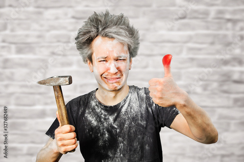 Dirty young builder guy fail is hold a hammer, hit the finger on brick wall background at home during repairs