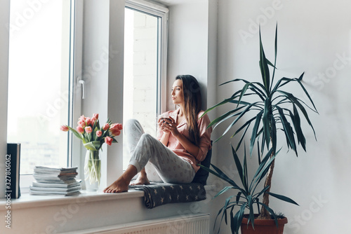 Thoughtful young woman in cozy pajamas looking through the window while resting at home