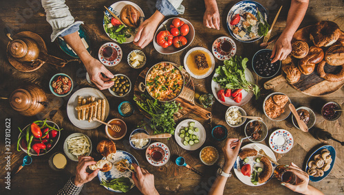 Turkish breakfast. Flat-lay of family eating pastry, vegetables, greens, cheeses, fried eggs, jams from oriental tableware, tea in copper pot and tulip glasses over rustic wooden background, top view