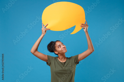 Pretty young african american woman girl in casual t-shirt posing isolated on bright blue background. People lifestyle concept. Mock up copy space. Hold yellow empty blank Say cloud, speech bubble.
