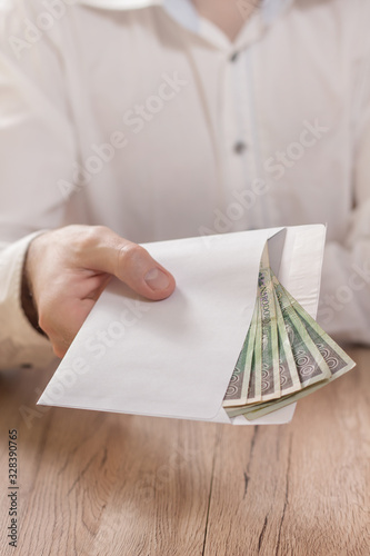 Męska dłoń trzyma białą kopertę wypełnioną banknotami polskiej waluty i podaje ją ponad stołem.